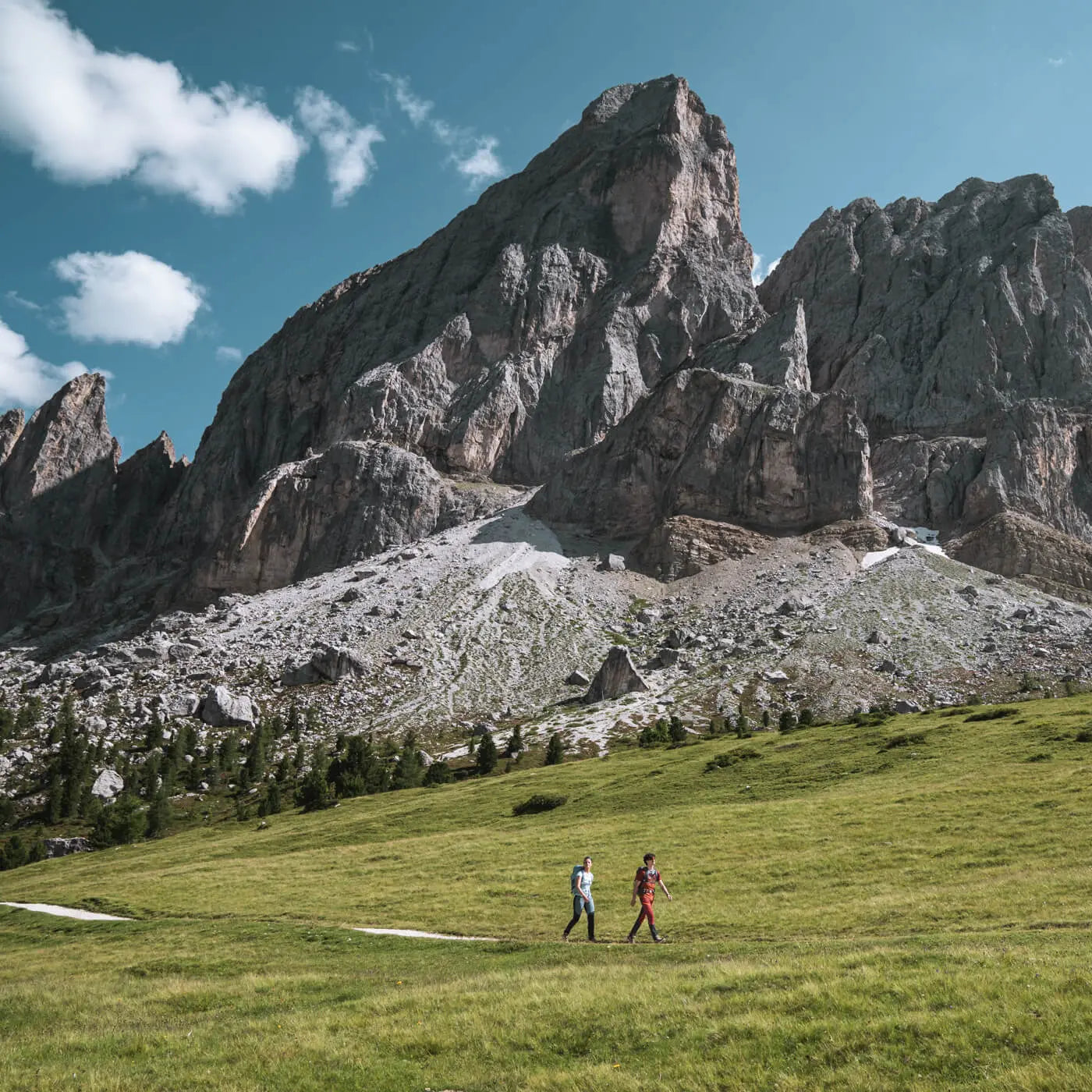 Two people walking on a grassy field with towering mountain peaks in the background.