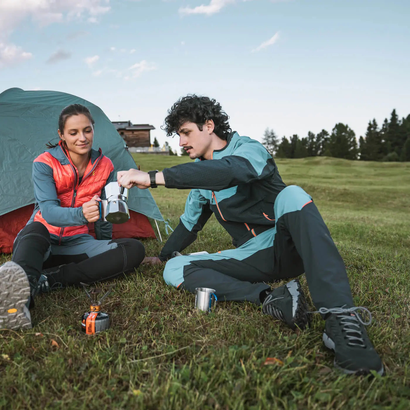 Two people camping outdoors, sitting near a tent and cooking on a portable stove.