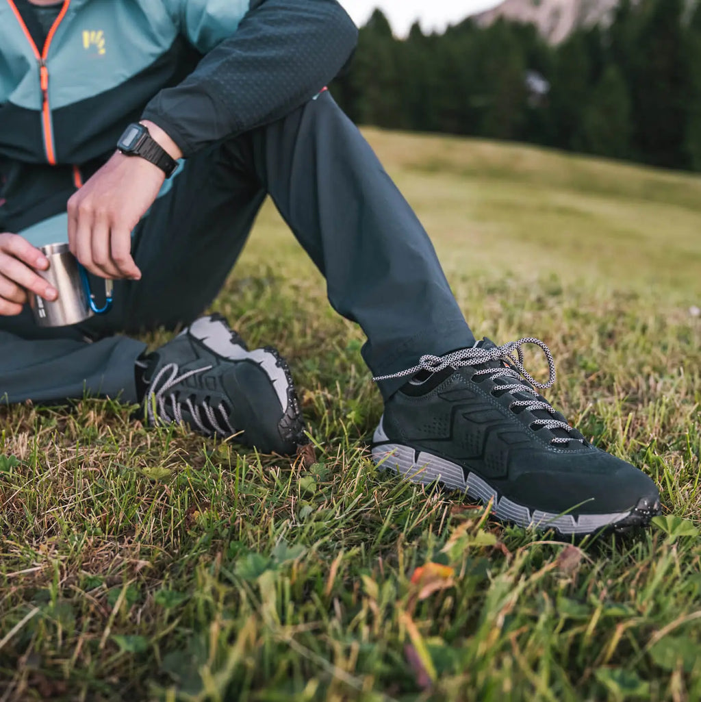 Person wearing black hiking shoes with white soles on grass, holding a metal cup.