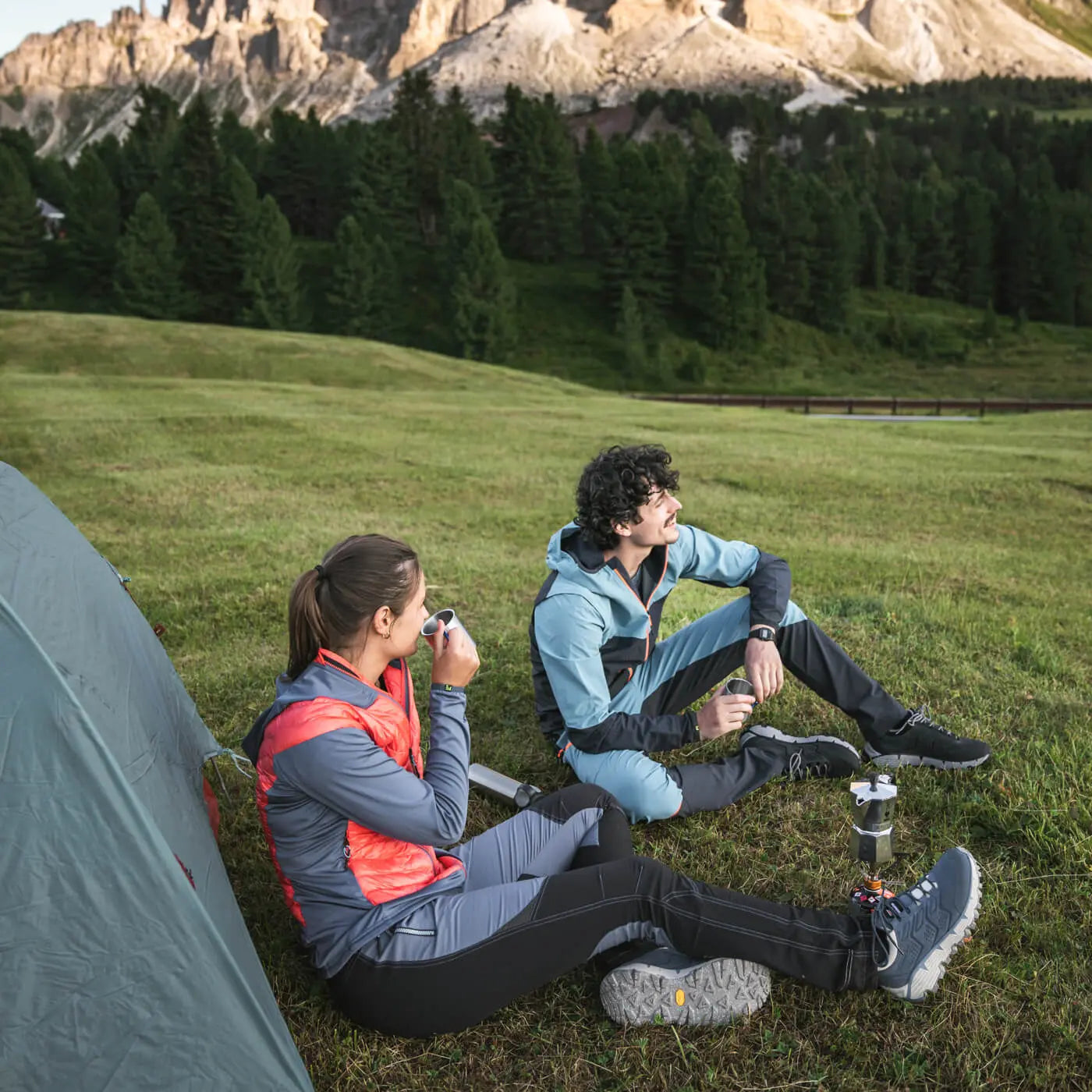 Two people sitting by a tent in a grassy field with mountains in the background