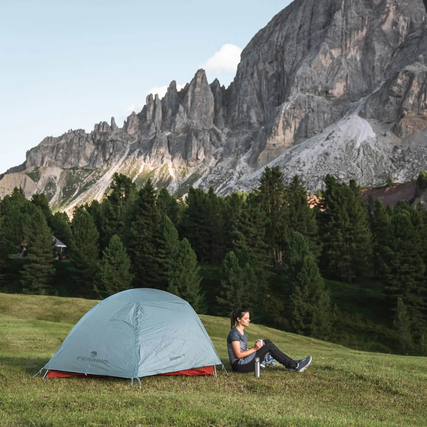 Person sitting next to a tent in a mountainous area with trees and cliffs.