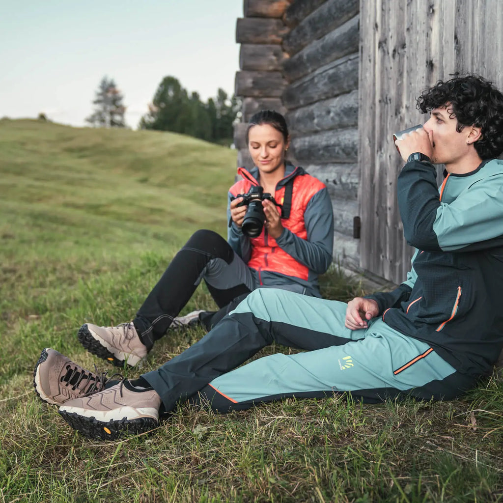 Two people sitting on grass near a wooden cabin, one taking a photo with a camera.