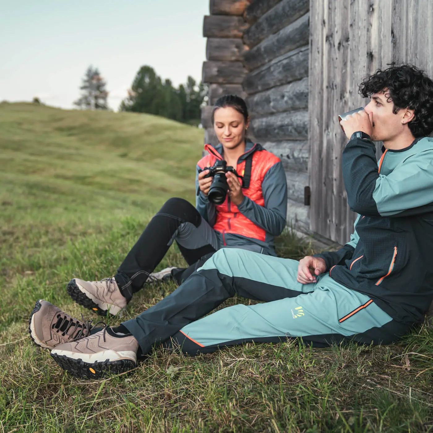 Two people sitting on grass near a wooden cabin, one taking a photo with a camera.