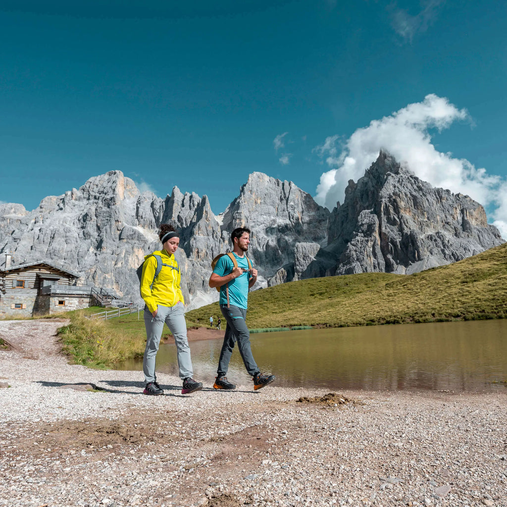 Two hikers walking near a mountain lake with a mountainous landscape in the background.