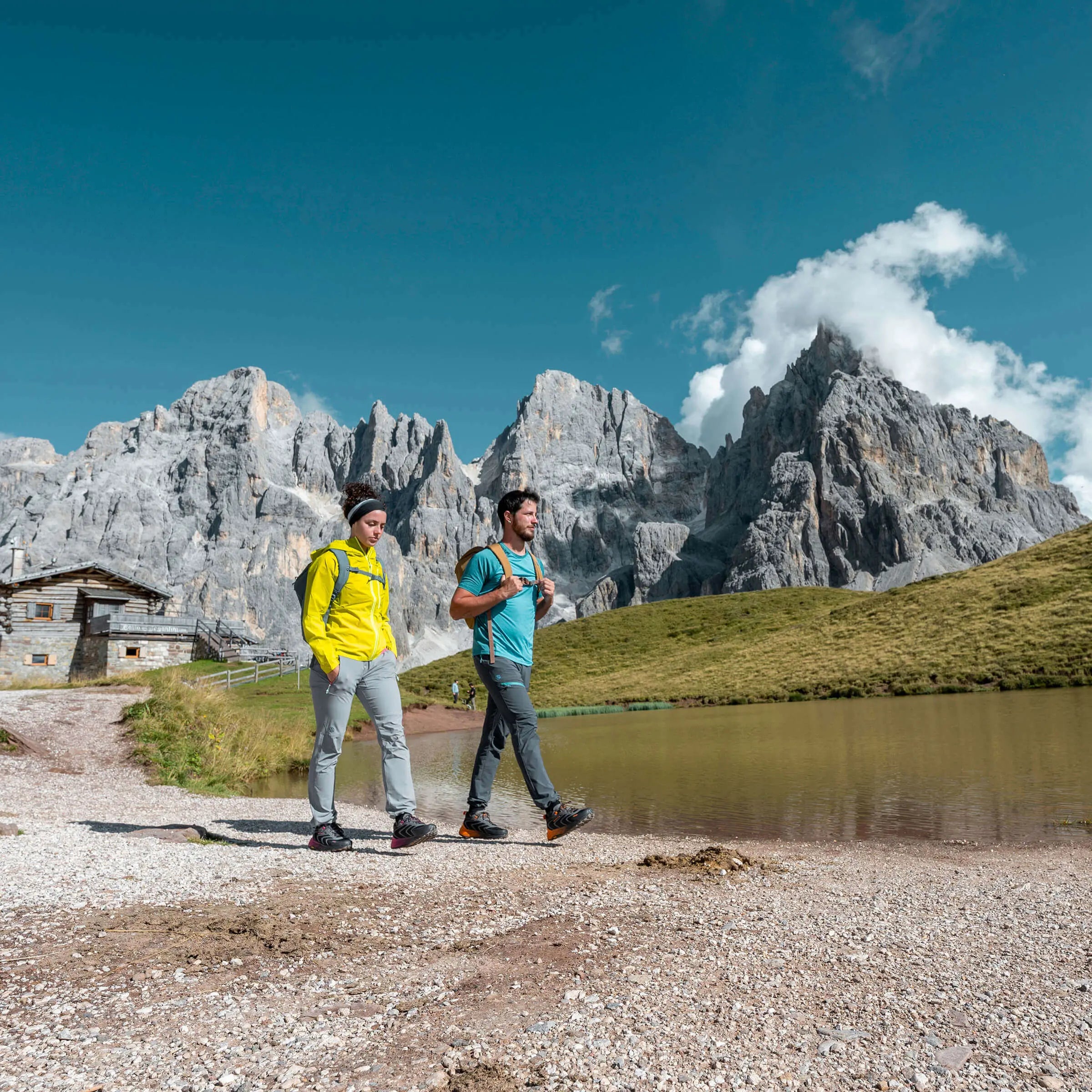 Two hikers walking near a mountain lake with a mountainous landscape in the background.