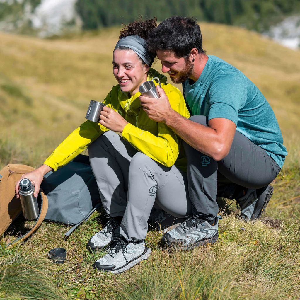 Two people sitting on a grassy hillside, enjoying a drink from thermos cups.