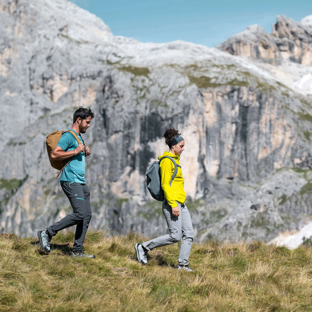 Two hikers with backpacks walking on a grassy hill with mountains in the background