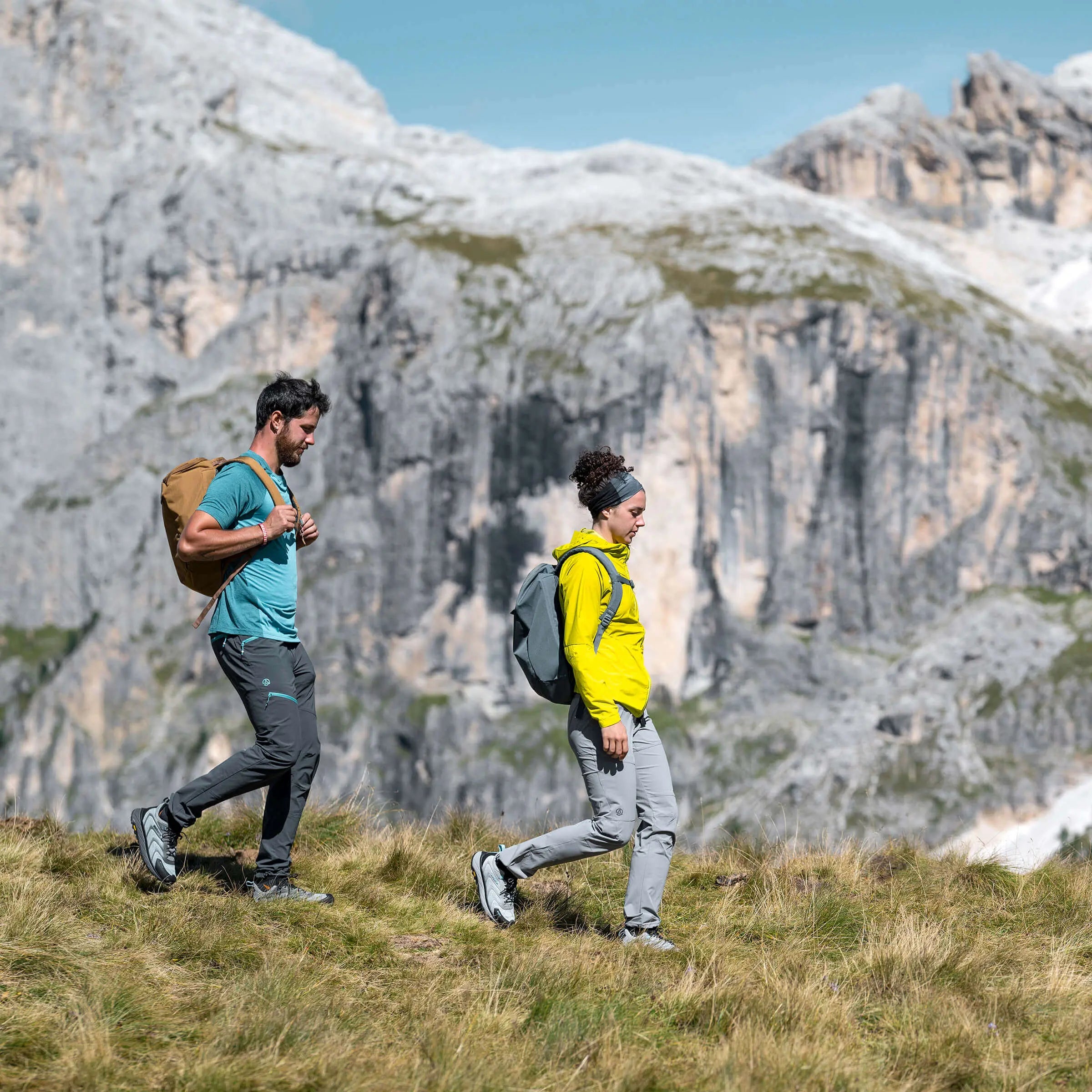 Two hikers with backpacks walking on a grassy hill with mountains in the background