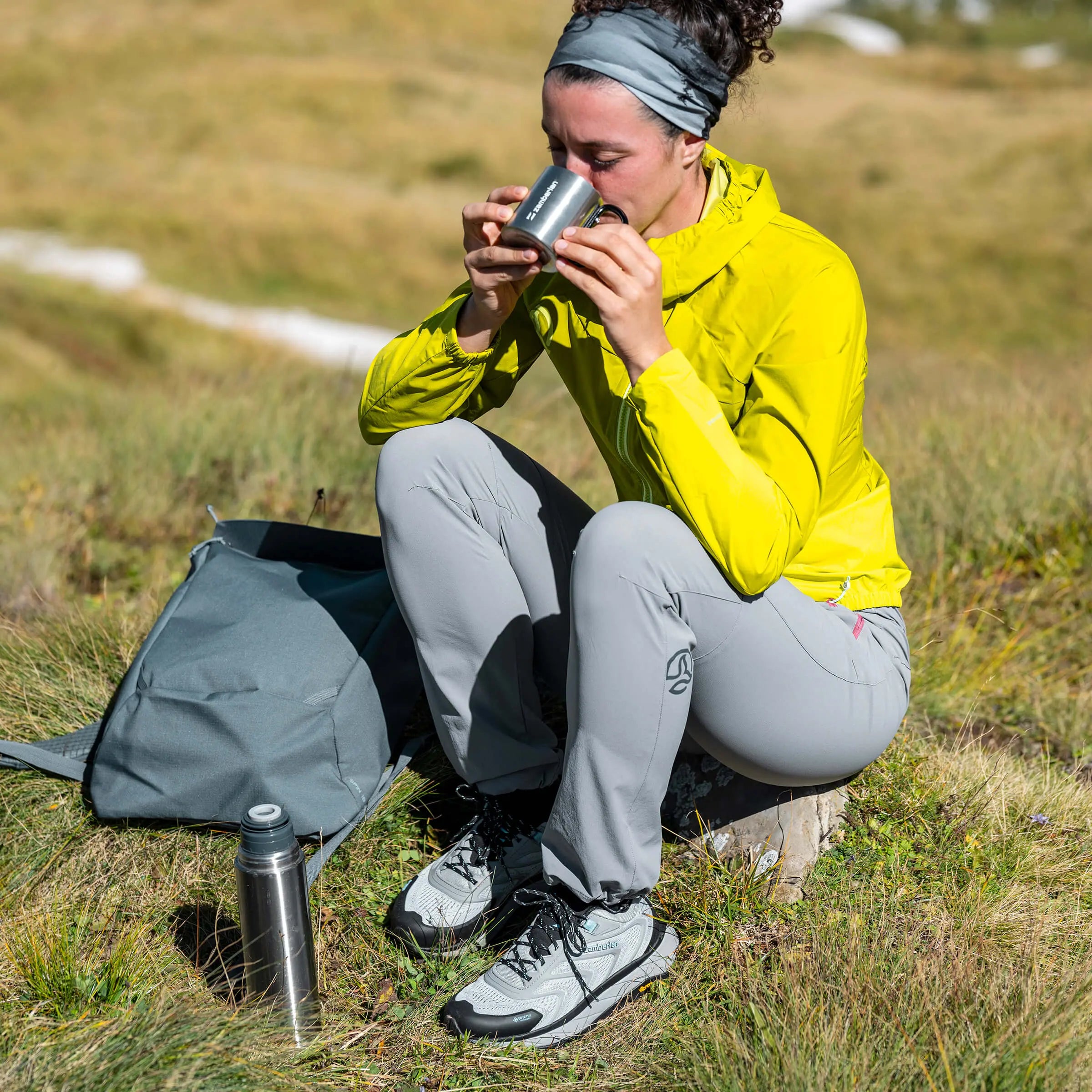 Person in yellow jacket and gray pants sitting on a log in a grassy field, drinking from a metal cup.