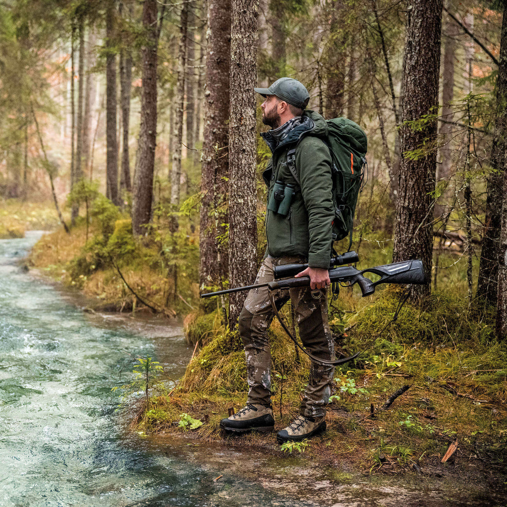Man in hunting gear walking along a forest path with a rifle.