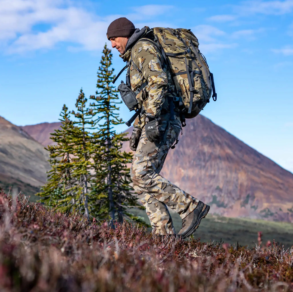 Person in camouflage gear with a backpack hiking in a mountainous landscape.