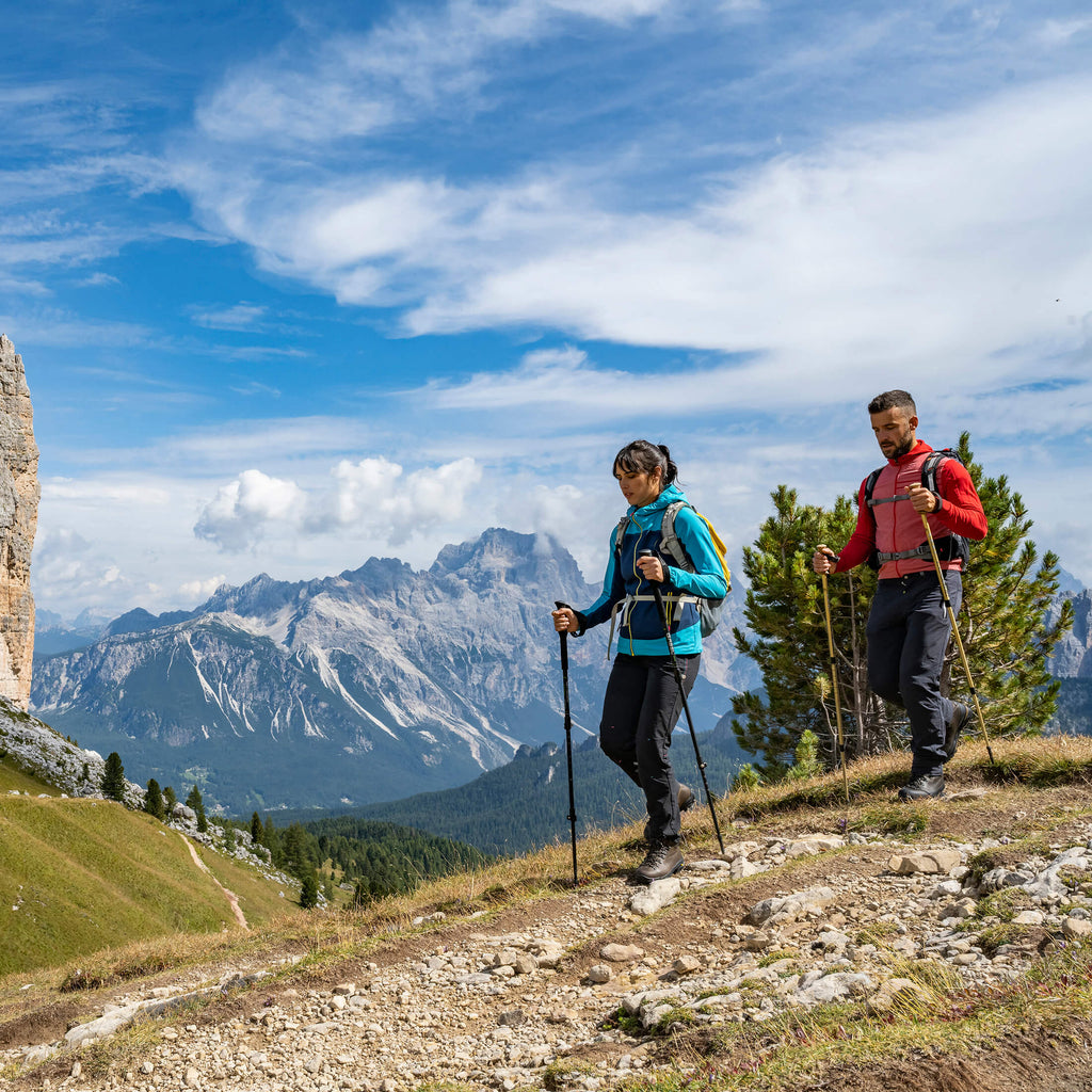 Two hikers on a mountain trail with a scenic view of mountains and blue sky.