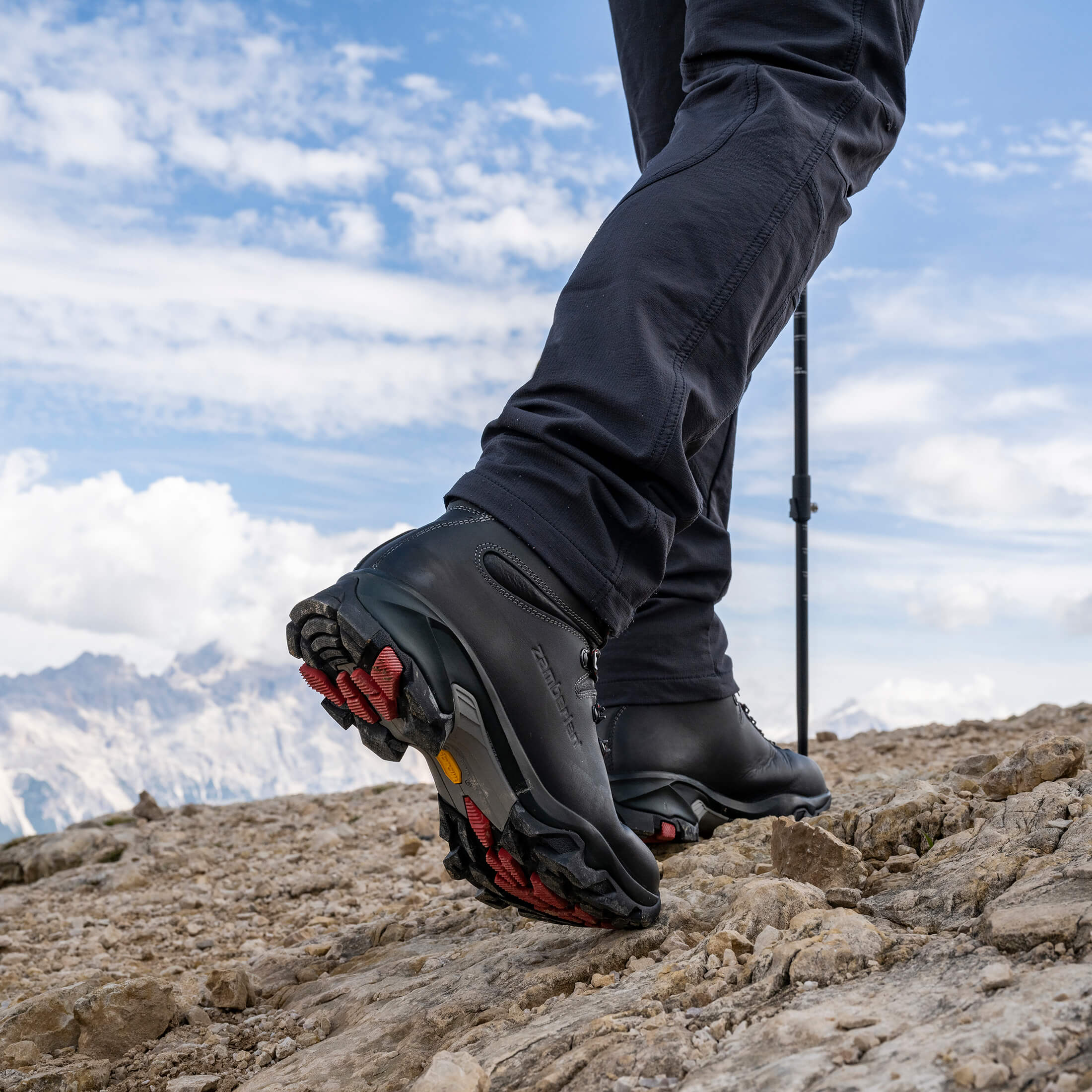 Person hiking on a mountain with black hiking boots and pants, against a blue sky.