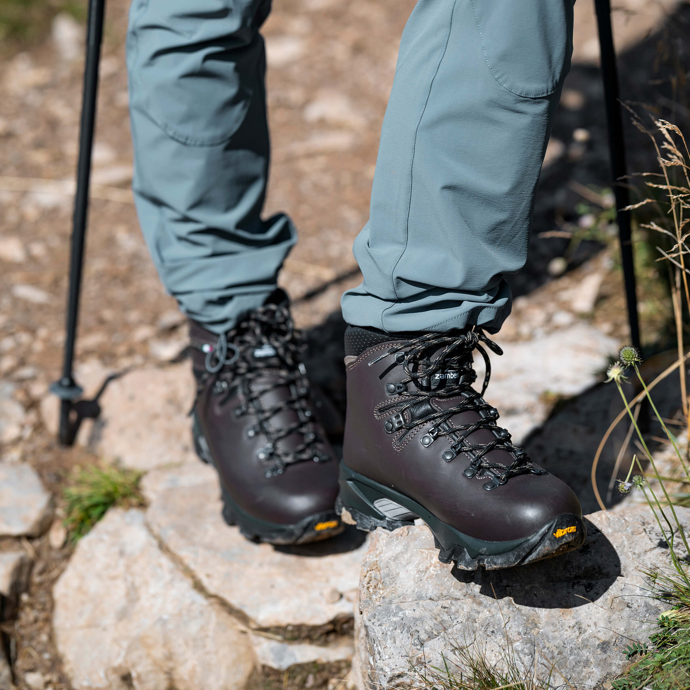 Hiking boots on rocky terrain with hiking poles and green pants.