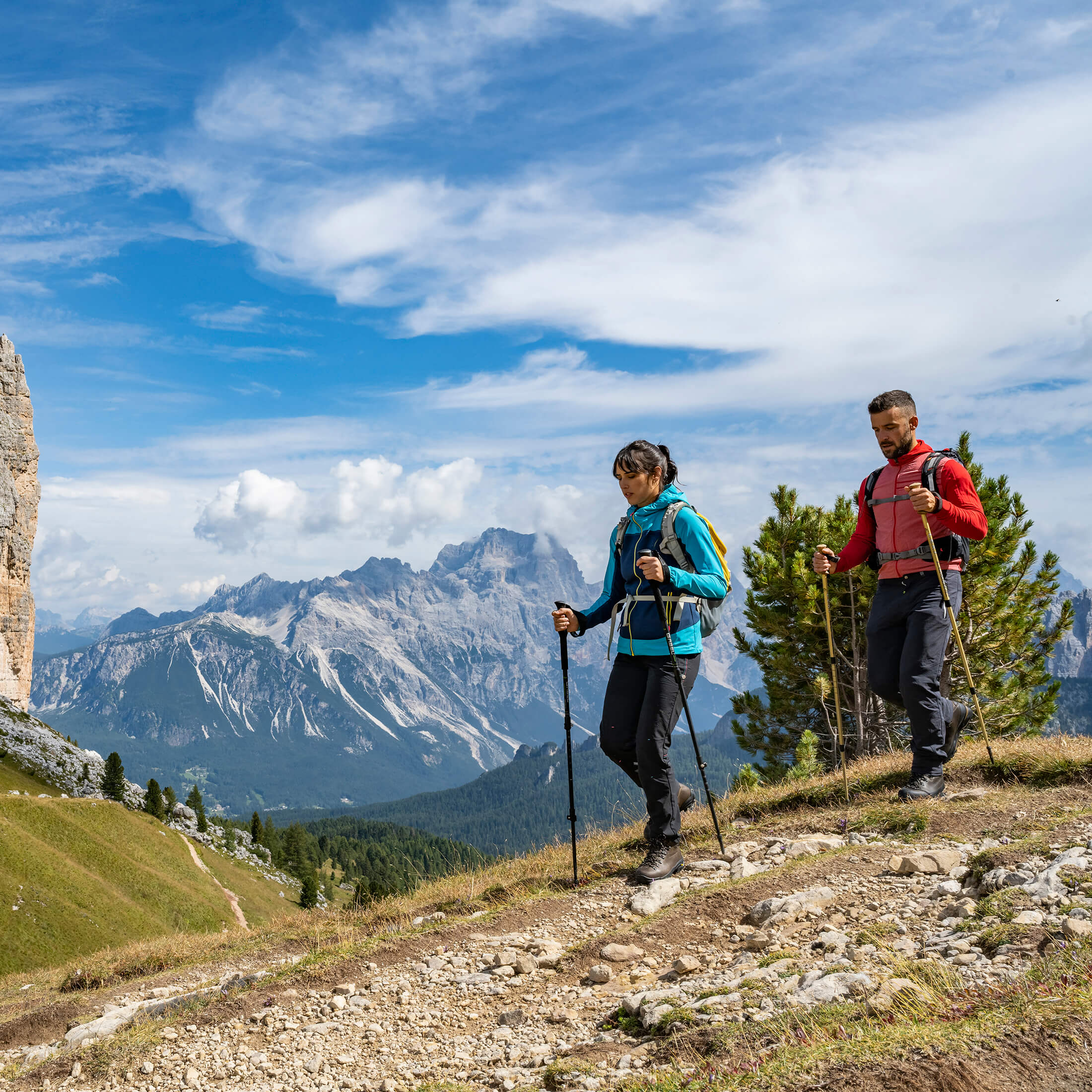Two hikers on a mountain trail with a scenic view of mountains and blue sky.