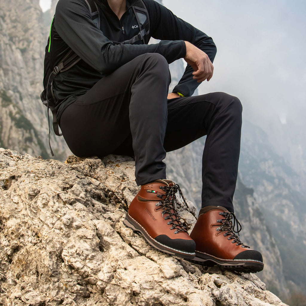Person wearing brown hiking boots sitting on a rocky mountain ledge with a mountainous background.