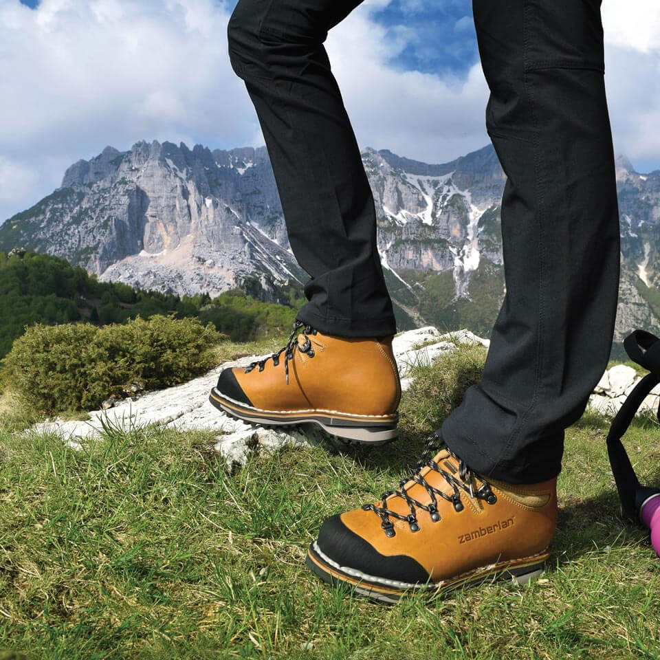Person wearing tan hiking boots with black soles in a mountainous landscape.