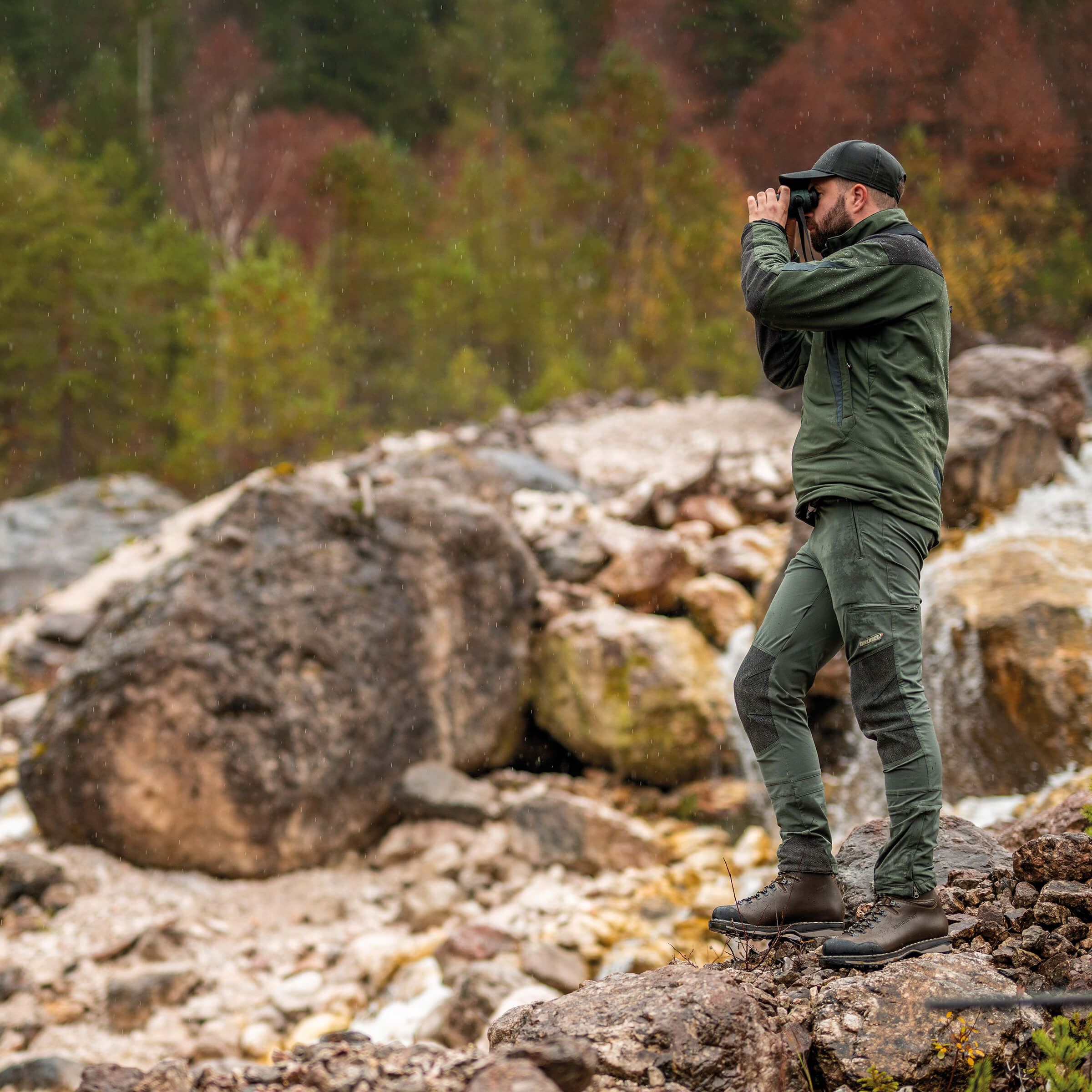 Man in green hiking gear using binoculars in a forested area with rocks and trees.