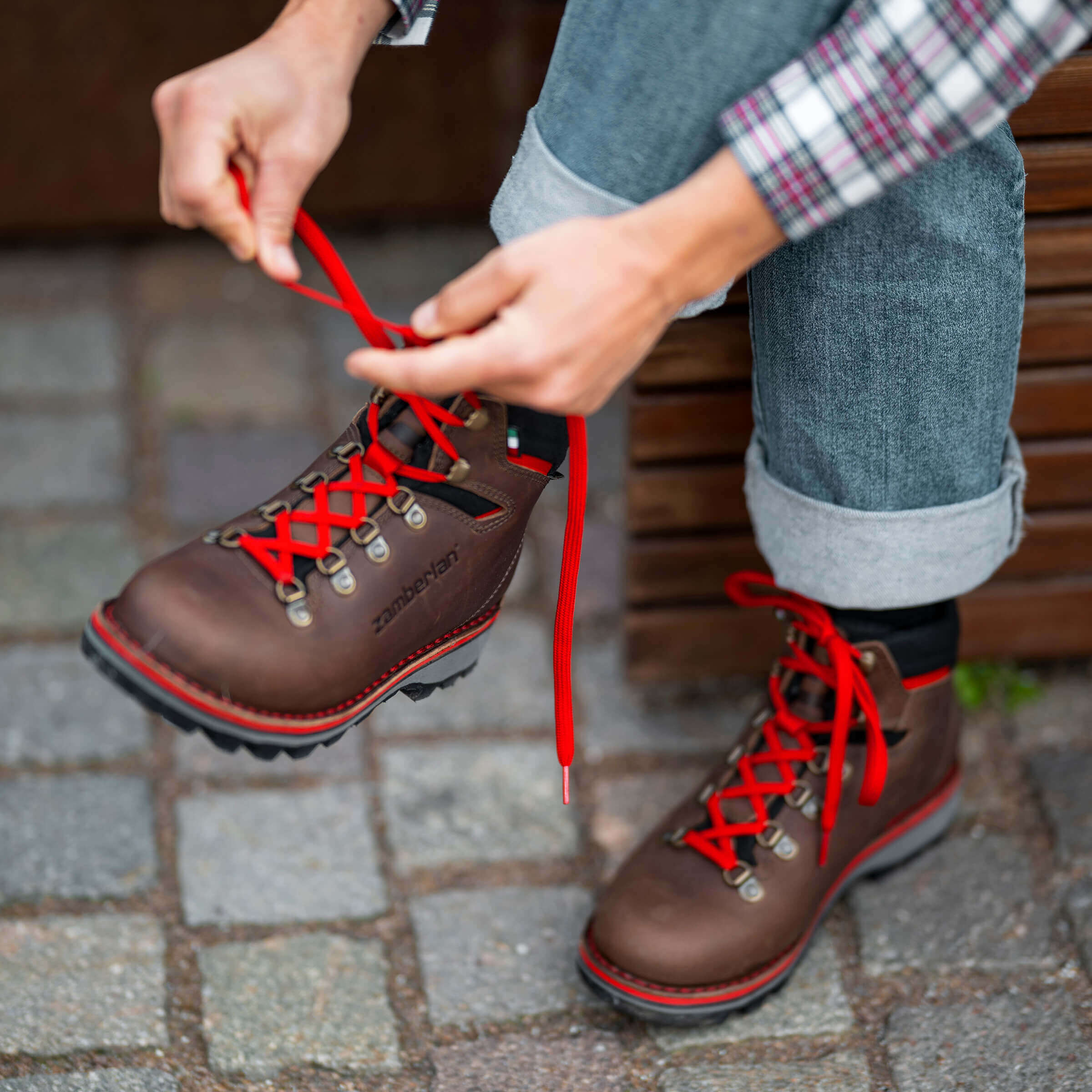 Person tying red laces on brown boots with a plaid shirt and jeans.
