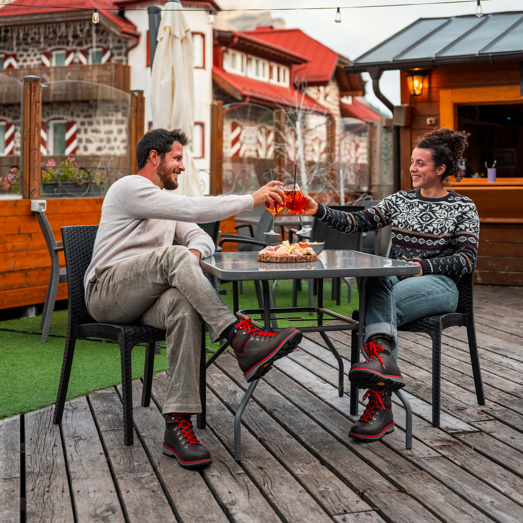 Two people sitting at a table outdoors, clinking glasses and smiling.