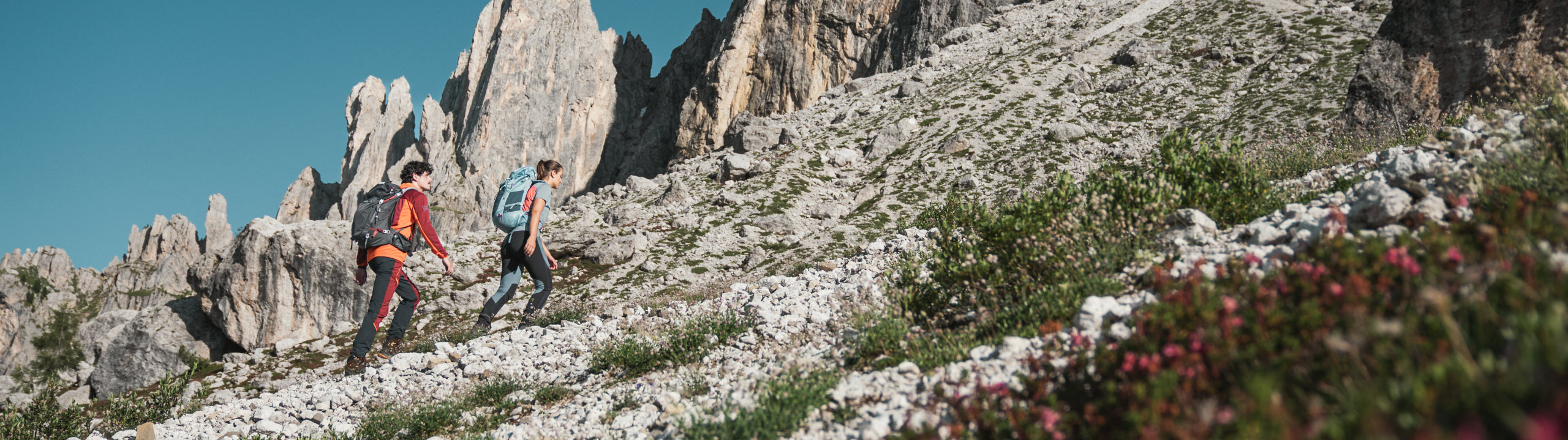 Two hikers on a mountain trail with rocky cliffs and clear sky.
