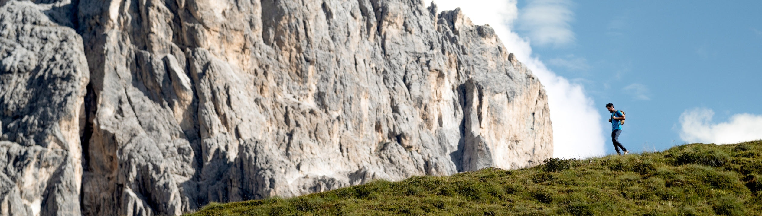 Person hiking on a grassy hill with a large rocky cliff and blue sky in the background
