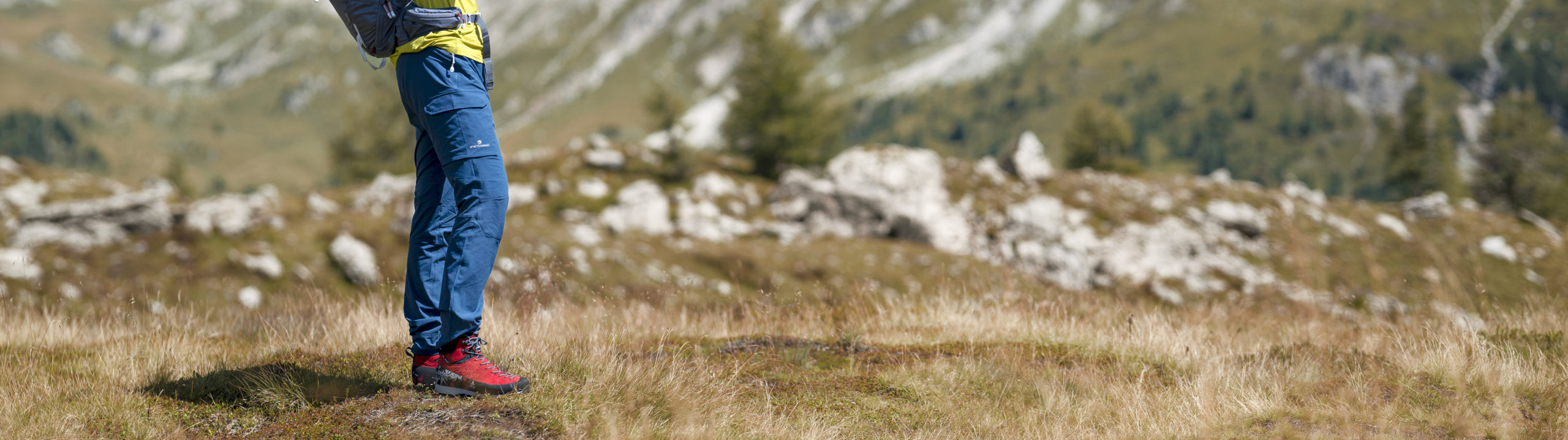 Person wearing blue pants and red shoes standing on a grassy hillside with mountains in the background