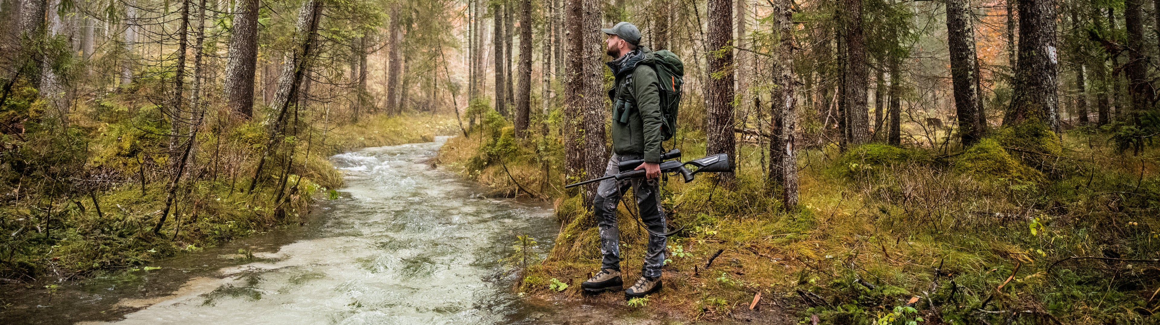 Hunter walking through a forest with a rifle, surrounded by trees and underbrush.