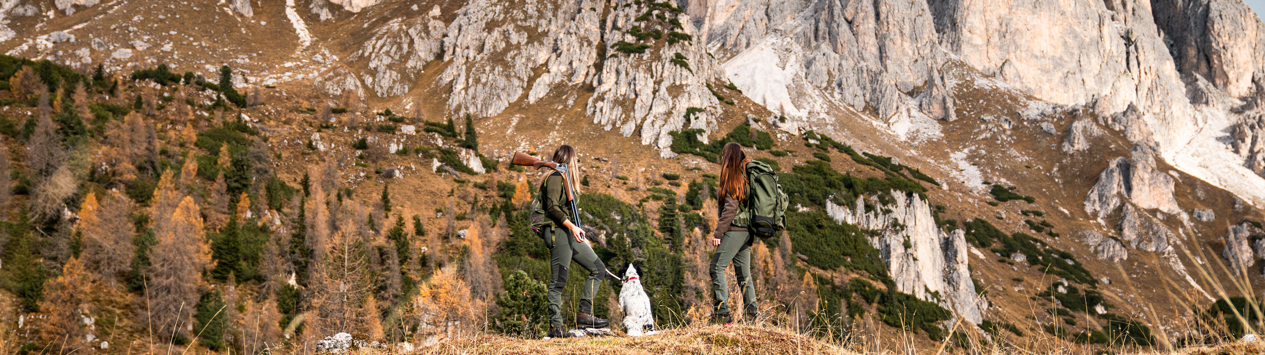 Two hikers with a dog on a mountain trail with rocky cliffs and trees in the background.