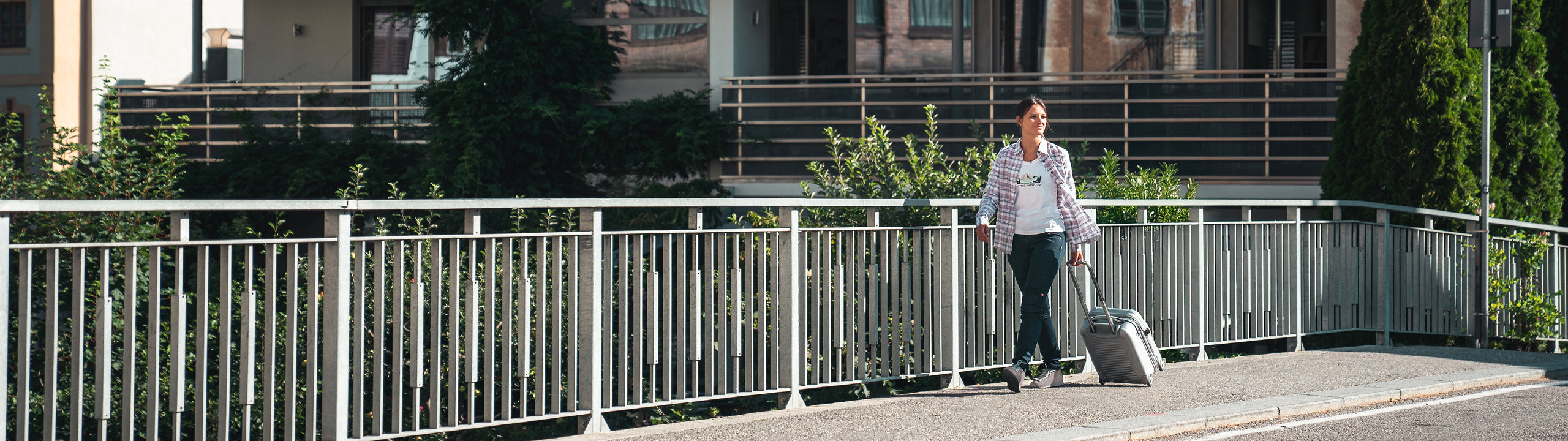 Person with a suitcase walking on a bridge with trees and buildings in the background